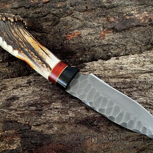 May include: A handcrafted knife with a textured blade and a handle made of antler, wood, and black accents. The blade has a unique pattern. The text "RZR BLADE" is visible near the bottom.