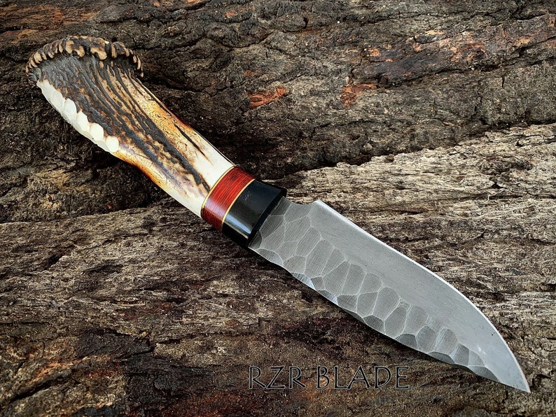 May include: A handcrafted knife with a textured blade and a handle made of antler, wood, and black accents. The blade has a unique pattern. The text "RZR BLADE" is visible near the bottom.