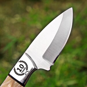 May include: A silver-colored metal knife blade with a sharp, curved edge. The bolster features a circular laurel wreath design surrounding the text 10 YEARS, with the word ANNIVERSARY engraved vertically along the side of the metal bolster.