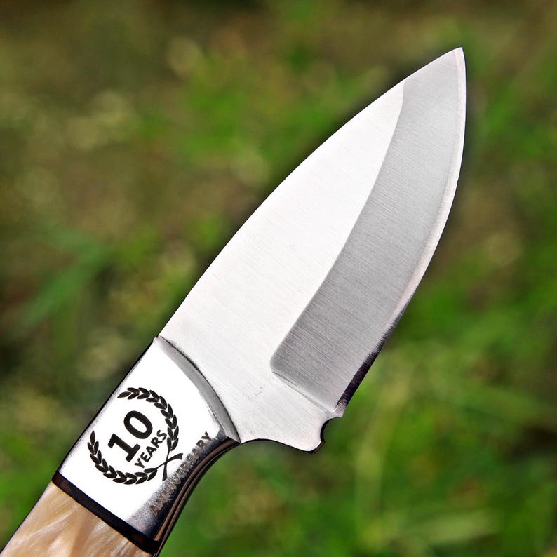 May include: A silver-colored metal knife blade with a sharp, curved edge. The bolster features a circular laurel wreath design surrounding the text 10 YEARS, with the word ANNIVERSARY engraved vertically along the side of the metal bolster.