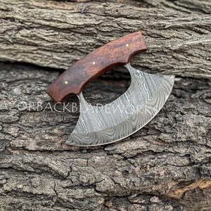 May include: A handcrafted Damascus steel ulu knife with a dark brown wood handle. The blade features a unique, swirling pattern.  The knife is displayed on a rustic, gray log background. Razorbackbladework is subtly visible on the blade. The handle has brass pins. This is a high-quality, hand-forged blade perfect for kitchen and other culinary tasks.
