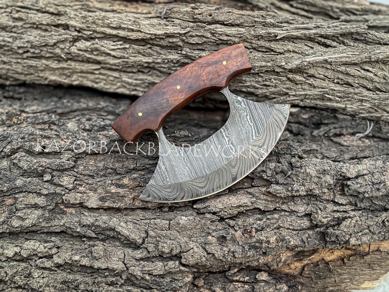 May include: A handcrafted Damascus steel ulu knife with a dark brown wood handle. The blade features a unique, swirling pattern.  The knife is displayed on a rustic, gray log background. Razorbackbladework is subtly visible on the blade. The handle has brass pins. This is a high-quality, hand-forged blade perfect for kitchen and other culinary tasks.