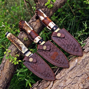 May include: Three fixed-blade knives with textured antler handles and dark wood accents, each featuring a small circular inlay with a red cross. The knives are housed in brown leather sheaths with basket-weave embossing and brass snap closures.