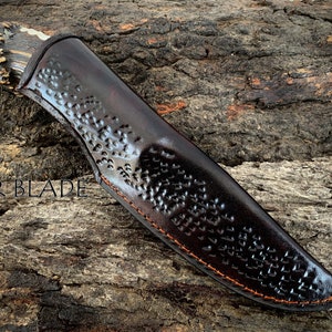 May include: A hunting knife with an antler handle and a dark brown leather sheath. The sheath features a geometric pattern and orange stitching. The text "RZR BLADE" is visible on the wood surface.