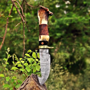 May include: A fixed-blade knife featuring a Damascus steel blade with a gut hook and a handle constructed from a combination of textured brown antler, smooth dark wood, and a black bolster with a small circular emblem featuring a red cross.
