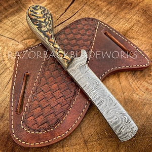 Puede incluir: Un cuchillo artesanal de acero Damasco con mango de madera con patr&oacute;n y funda de cuero. La funda tiene un patr&oacute;n tejido y est&aacute; cosida con hilo marr&oacute;n. El cuchillo est&aacute; apoyado sobre una superficie de madera.