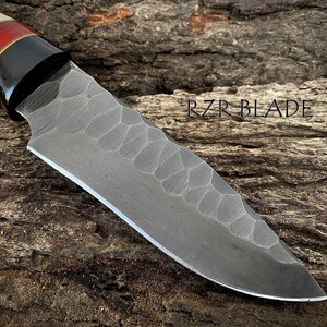 May include: A handcrafted knife with a textured blade and a handle featuring bone, red, and black accents. The blade has a unique pattern and the text "RZR BLADE" is visible.