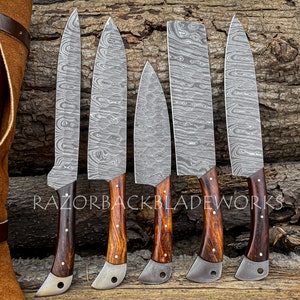 May include: Set of six kitchen knives with wooden handles and a Damascus steel blade pattern. The knives are arranged on a wooden surface with a leather knife roll in the background. The text "RAZORBACKBLADEWORKS" is visible on the surface.