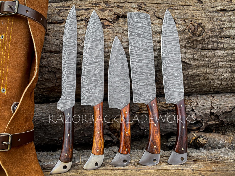 May include: Set of six kitchen knives with wooden handles and a Damascus steel blade pattern. The knives are arranged on a wooden surface with a leather knife roll in the background. The text "RAZORBACKBLADEWORKS" is visible on the surface.