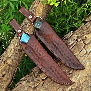 May include: Two knives with patterned brown leather sheaths featuring embossed basket-weave textures and stitching. One knife handle is bright blue, while the other is green with a textured pattern. Each sheath has a strap with a metal snap.
