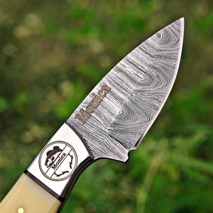May include: A fixed-blade knife with a Damascus steel blade featuring a distinct wavy pattern and the name Monica engraved on the metal. The bolster displays a circular logo with a fish illustration and the name Monica, paired with a cream handle.
