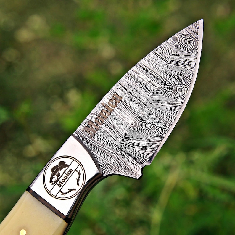 May include: A fixed-blade knife with a Damascus steel blade featuring a distinct wavy pattern and the name Monica engraved on the metal. The bolster displays a circular logo with a fish illustration and the name Monica, paired with a cream handle.