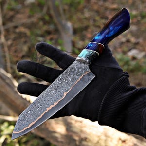 May include: A fixed-blade knife featuring a patterned Damascus steel blade with a copper-colored edge. The handle is crafted from a polished, deep blue resin material with swirling patterns, accented by a turquoise and black marbled bolster section.