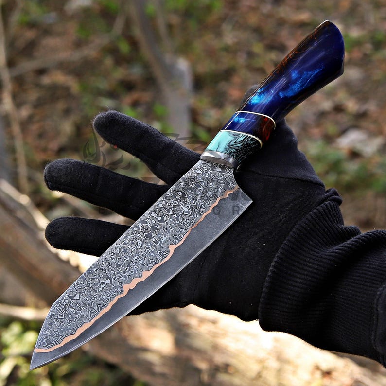May include: A fixed-blade knife featuring a patterned Damascus steel blade with a copper-colored edge. The handle is crafted from a polished, deep blue resin material with swirling patterns, accented by a turquoise and black marbled bolster section.