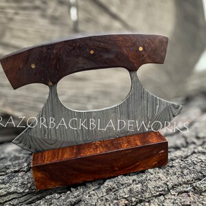 May include: A handcrafted Damascus steel ulu knife with a curved blade and a dark brown wood handle. The knife features a unique pattern on the blade and is displayed on a matching wooden stand.  The brand name 'RAZORBACKBLADEWORKS' is subtly visible.  The handle is accented with small gold colored pins. This high-quality, artisan knife is perfect for any kitchen or collection.