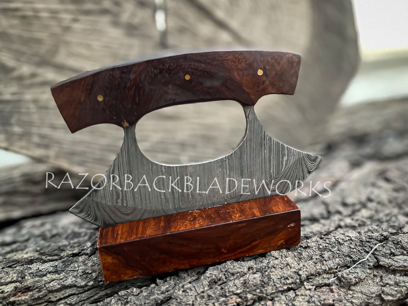 May include: A handcrafted Damascus steel ulu knife with a curved blade and a dark brown wood handle. The knife features a unique pattern on the blade and is displayed on a matching wooden stand.  The brand name 'RAZORBACKBLADEWORKS' is subtly visible.  The handle is accented with small gold colored pins. This high-quality, artisan knife is perfect for any kitchen or collection.