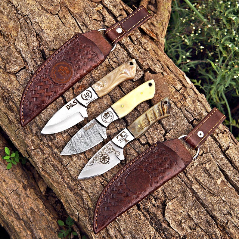 May include: Three fixed-blade knives with distinct handles and two brown leather sheaths with basket-weave patterns. The top knife has a pearlescent handle and text reading D&S 10th Anniversary. The middle knife has a cream handle and a Damascus steel blade. The bottom knife has a textured handle and a blade engraved with a compass design and the name Tyler.