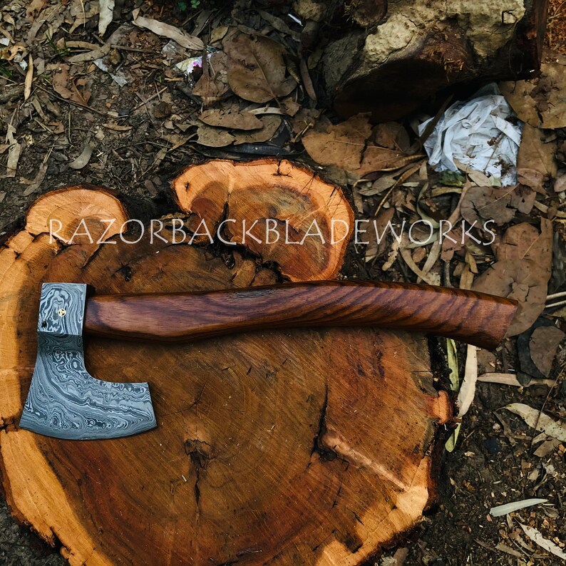 May include: A hand-forged axe with a Damascus steel blade and a wooden handle. The axe is resting on a wooden stump. The text "RAZORBACKBLADEWORKS" is visible on the stump.