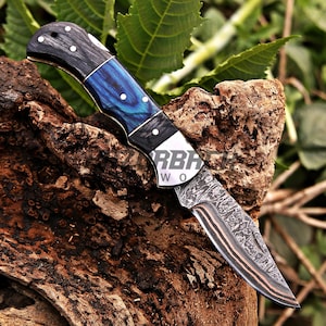 Op de afbeelding: Een inklapbaar zakmes met een Damascus stalen lemmet met zichtbare gelaagde patronen en koperen accenten. Het handvat is gemaakt van zwart en blauw gemarmerd materiaal, vastgezet met zilveren pinnen en een metalen bolster met tekst.