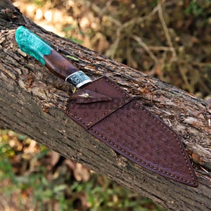 May include: A fixed-blade knife featuring a handle made of dark wood and translucent emerald green resin, paired with a brown leather sheath. The sheath displays a stamped basket-weave pattern, visible stitching, and a single brass-colored metal rivet.
