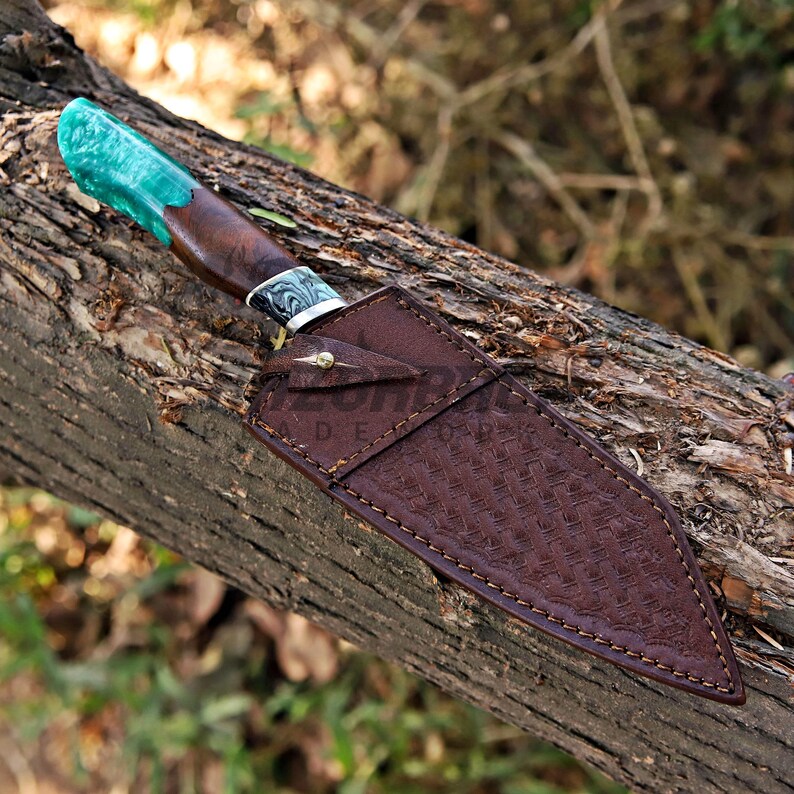 May include: A fixed-blade knife featuring a handle made of dark wood and translucent emerald green resin, paired with a brown leather sheath. The sheath displays a stamped basket-weave pattern, visible stitching, and a single brass-colored metal rivet.