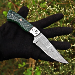 May include: A fixed-blade knife featuring a Damascus steel blade with a distinct wavy pattern. The handle is green with a dark honeycomb grid design, secured by brass pins. The bolster is engraved with the letters SB and a small antler icon.