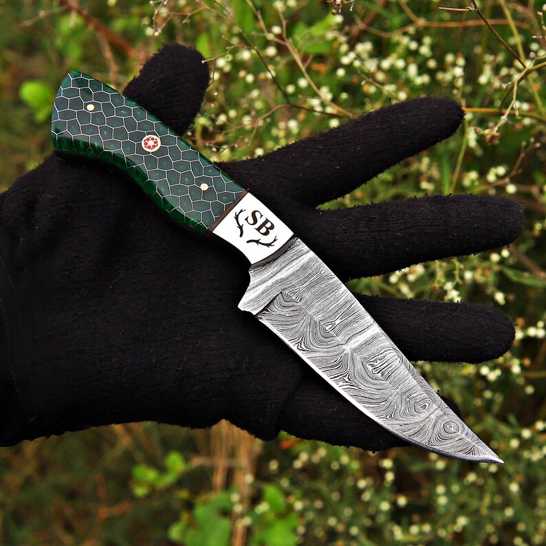 May include: A fixed-blade knife featuring a Damascus steel blade with a distinct wavy pattern. The handle is green with a dark honeycomb grid design, secured by brass pins. The bolster is engraved with the letters SB and a small antler icon.