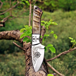 May include: A fixed-blade knife with a polished metal blade featuring an engraved compass rose and topographic lines. The blade is marked with the name TYLER. The handle is made of dark, textured wood with two brass pins and a lanyard hole.