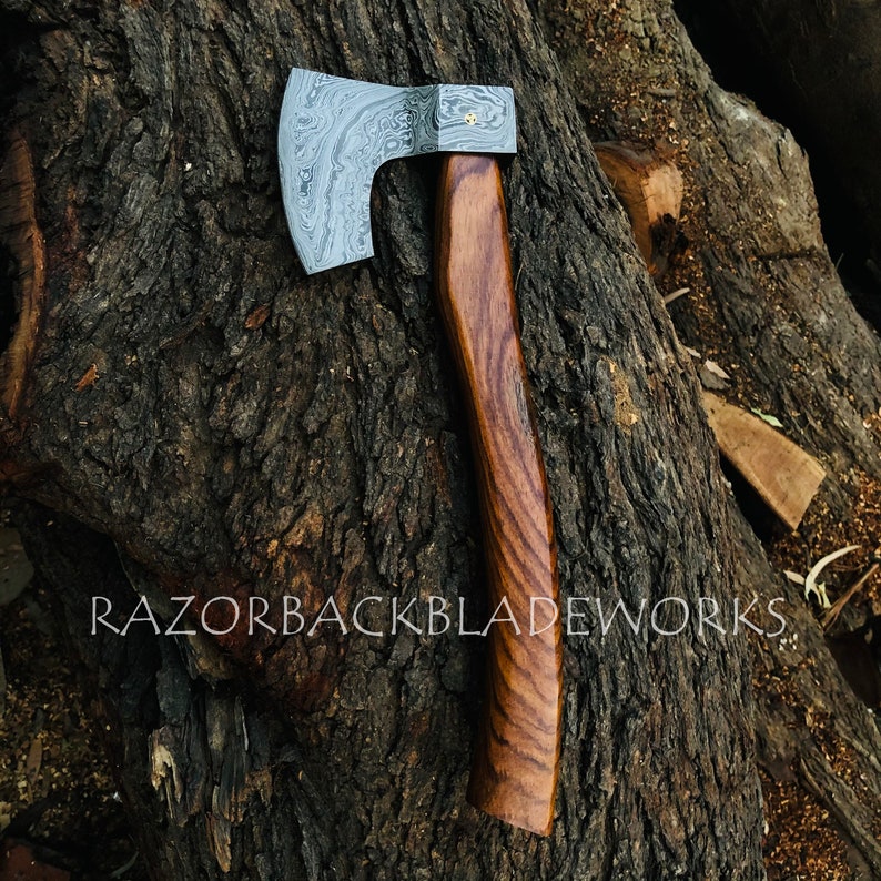 May include: A hand-forged axe with a Damascus steel blade and a wooden handle. The axe has a unique pattern on the blade and is resting on a piece of wood.