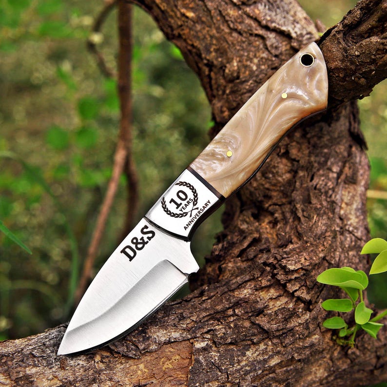 May include: A fixed-blade knife with a silver-toned metal blade featuring the text D&S and a 10 Years Anniversary emblem. The handle is made of a smooth, marbled beige and cream material with two small brass pins and a lanyard hole at the end.