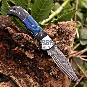 May include: A folding pocket knife featuring a Damascus steel blade with copper layers, a handle with black and blue marbled sections, and silver pins. The metal bolster is engraved with the partial text RBAC WO.