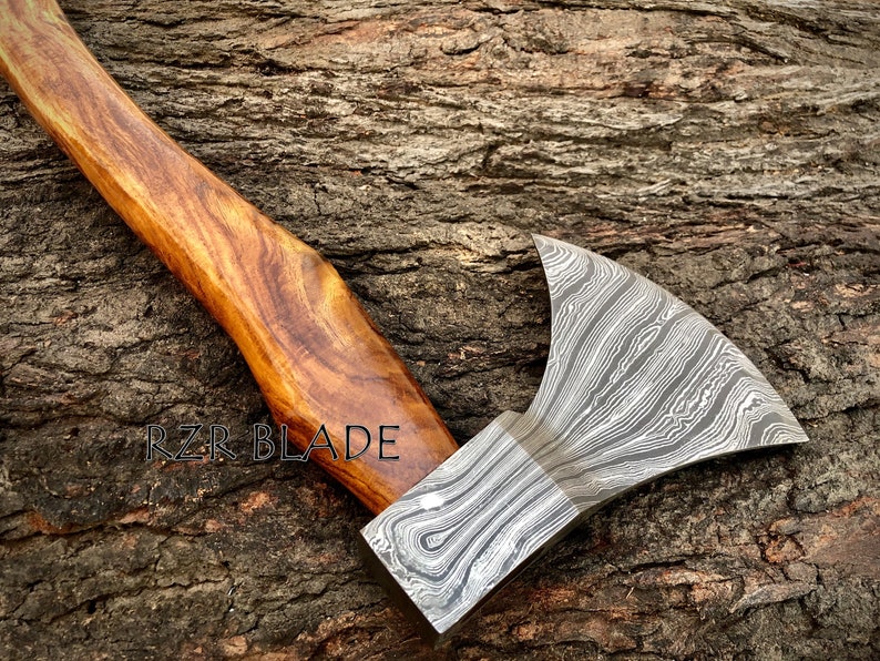 May include: A hand-forged axe with a wood handle. The axe head has a Damascus steel pattern and the handle is made of dark wood. The axe is resting on a wooden surface. The text "RZR BLADE" is visible on the handle.