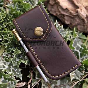 May include: A dark brown leather pouch with a brass snap button closure and yellow stitching. A small metal rod with a brass-colored tip is held in a side loop. The text RAZORBACK LEATHERWORKS is printed on the front of the leather pouch.