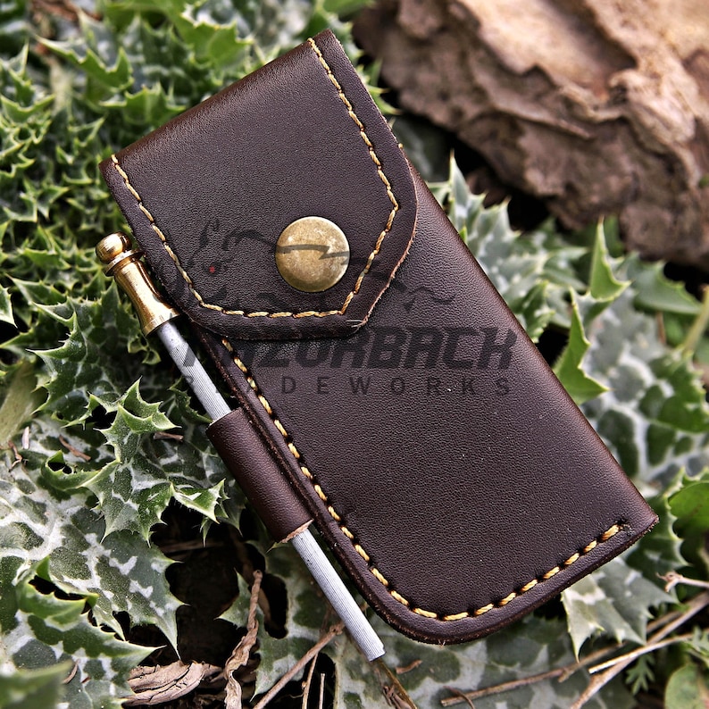 May include: A dark brown leather pouch with a brass snap button closure and yellow stitching. A small metal rod with a brass-colored tip is held in a side loop. The text RAZORBACK LEATHERWORKS is printed on the front of the leather pouch.