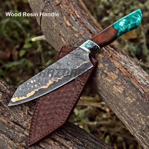 May include: A knife with a patterned Damascus steel blade and a handle made of dark wood and marbled green resin. The knife is accompanied by a brown leather sheath with a textured surface. The text Wood Resin Handle is visible.