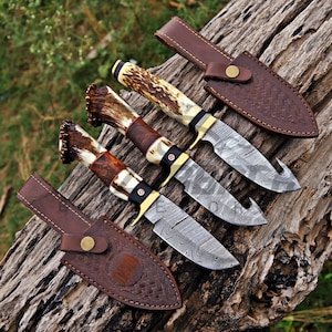Three handcrafted Damascus steel hunting knives with genuine antler handles and brass guards displayed on rustic tree bark in an outdoor setting.