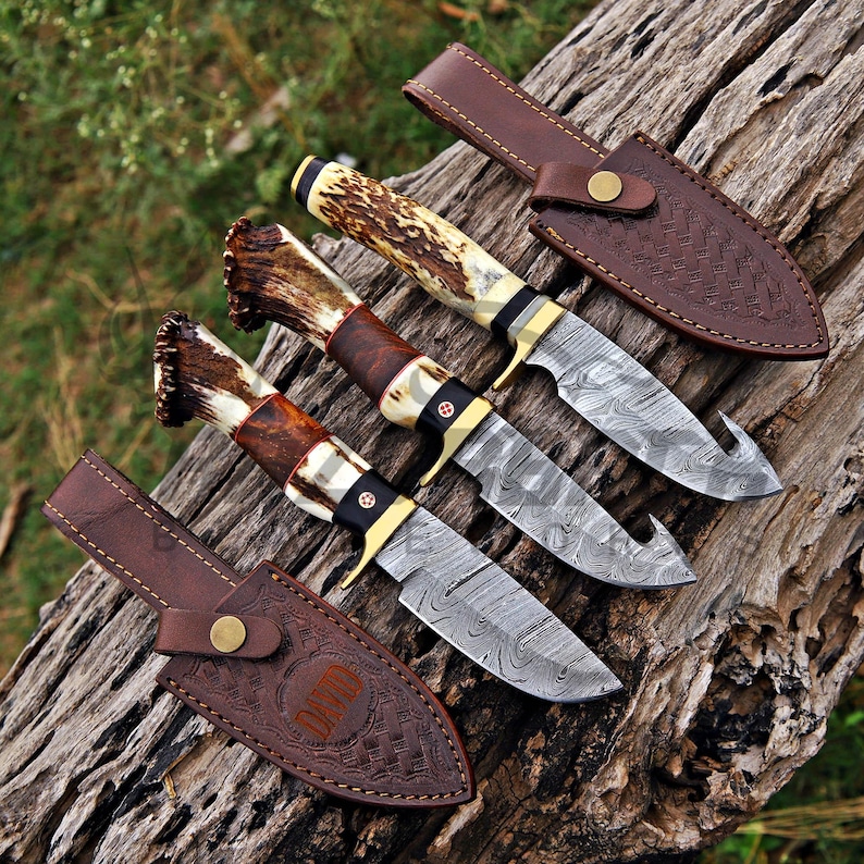 Three handcrafted Damascus steel hunting knives with genuine antler handles and brass guards displayed on rustic tree bark in an outdoor setting.