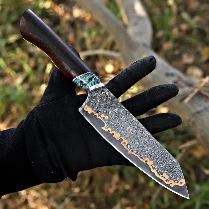 May include: A fixed-blade knife featuring a dark brown wooden handle, a decorative teal and silver bolster, and a patterned Damascus steel blade with a prominent copper-colored wavy line running along the edge of the metal surface.
