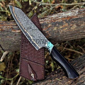 May include: A fixed-blade knife featuring a patterned Damascus steel blade with a copper-colored spine, a black handle with a turquoise inlay, and a brown leather sheath with a woven texture and a single metal stud fastener.