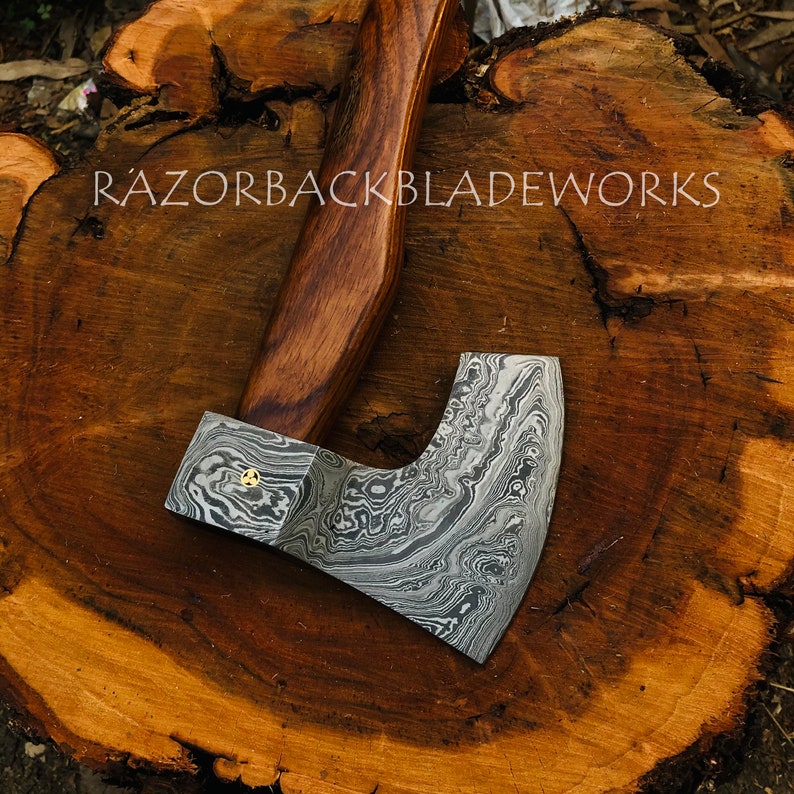 May include: A hand-forged axe with a wood handle and a damascus steel blade. The blade has a unique pattern and is secured with a brass rivet. The axe is resting on a wooden surface. The text "RAZORBACKBLADEWORKS" is visible in the background.