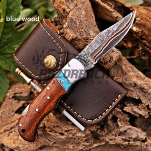 May include: A folding pocket knife with a patterned Damascus steel blade, a wooden handle, and a turquoise inlay. The knife is positioned on a brown leather sheath with yellow stitching and a brass snap button. The text blue wood is visible.