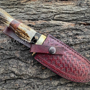 May include: A fixed-blade knife with a textured antler handle, a dark wood spacer, and a brass bolster, featuring a decorative mosaic pin. The knife is housed in a deep red, basket-weave embossed leather sheath with a metal snap closure.