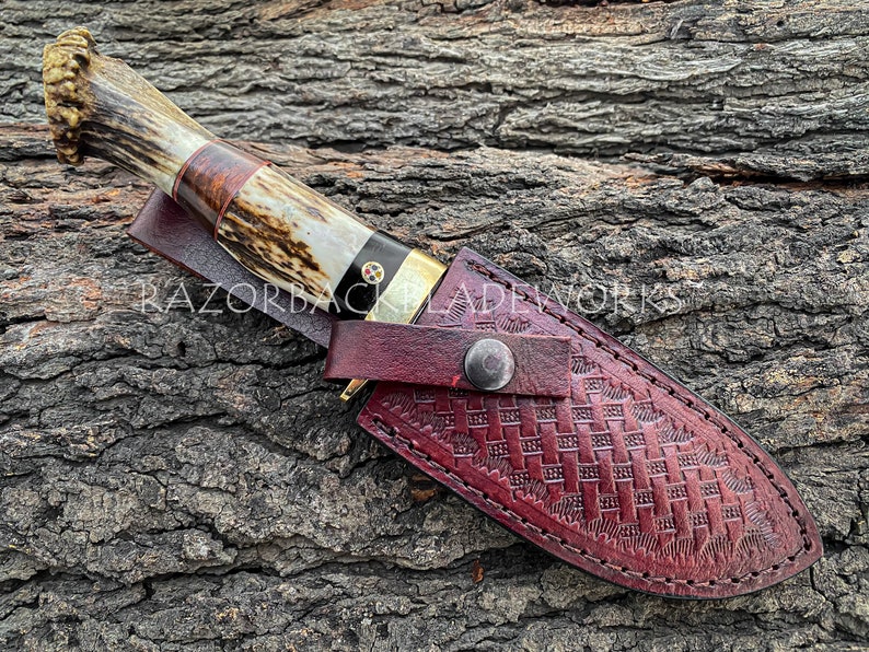 May include: A fixed-blade knife with a textured antler handle, a dark wood spacer, and a brass bolster, featuring a decorative mosaic pin. The knife is housed in a deep red, basket-weave embossed leather sheath with a metal snap closure.
