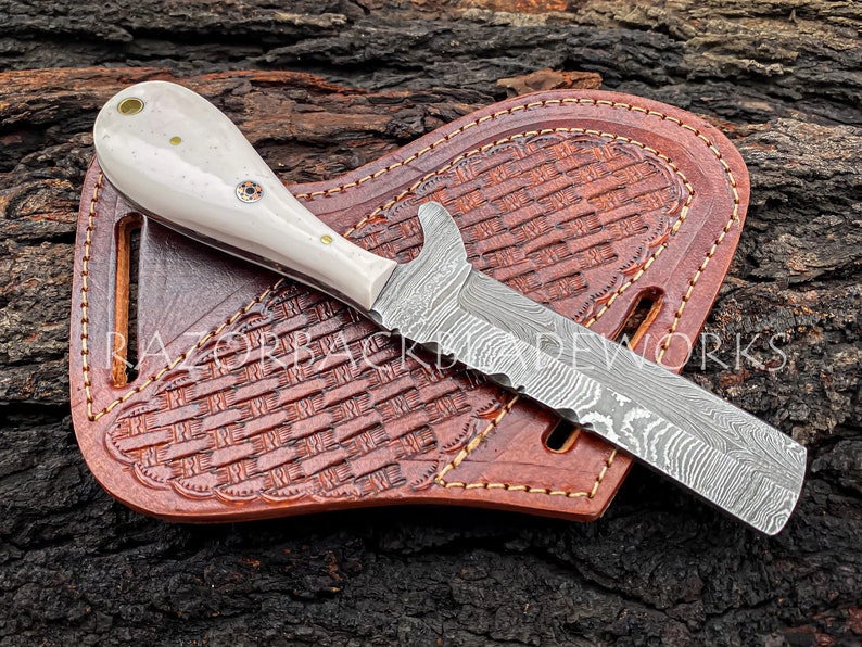 Puede incluir: Un cuchillo con mango blanco y hoja de acero damasco, descansando en una funda de cuero marr&oacute;n con un patr&oacute;n tejido. La funda tiene costuras decorativas alrededor de los bordes.