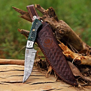 May include: A fixed-blade knife with a Damascus steel blade featuring a wavy pattern and the letters SB. The handle is green with a white honeycomb pattern and brass pins. It includes a brown leather sheath with a basket-weave texture and SB logo.