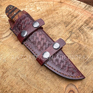 May include: A fixed-blade knife with a wood handle featuring a brass pin, housed in a dark burgundy leather sheath with a basket-weave pattern, secured by two metal snap buttons. The text RAZORBACKBLADEWORKS is visible across the sheath.