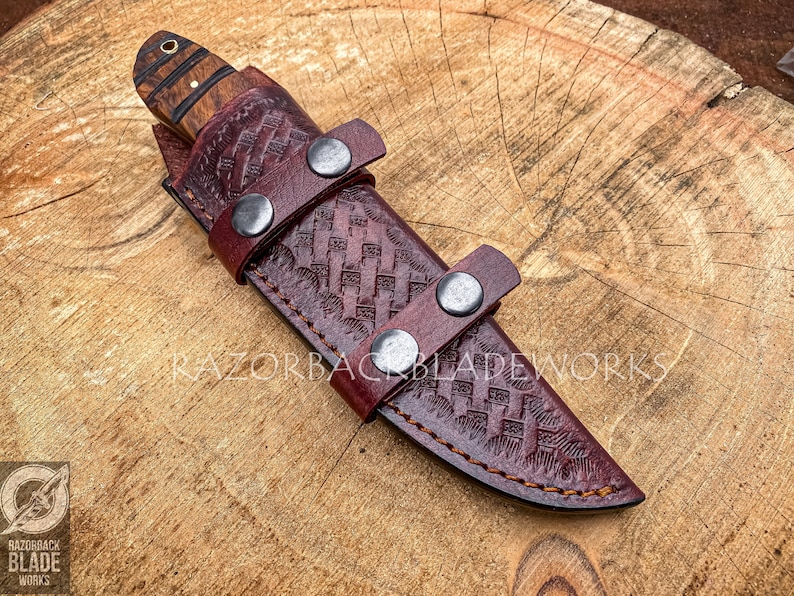 May include: A fixed-blade knife with a wood handle featuring a brass pin, housed in a dark burgundy leather sheath with a basket-weave pattern, secured by two metal snap buttons. The text RAZORBACKBLADEWORKS is visible across the sheath.