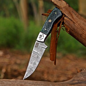 May include: A fixed-blade knife with a Damascus steel blade featuring a wavy pattern and the letters SB engraved. The handle is dark green with a black honeycomb pattern, gold-colored pins, and a bolster marked with the letters SB and antler icons.
