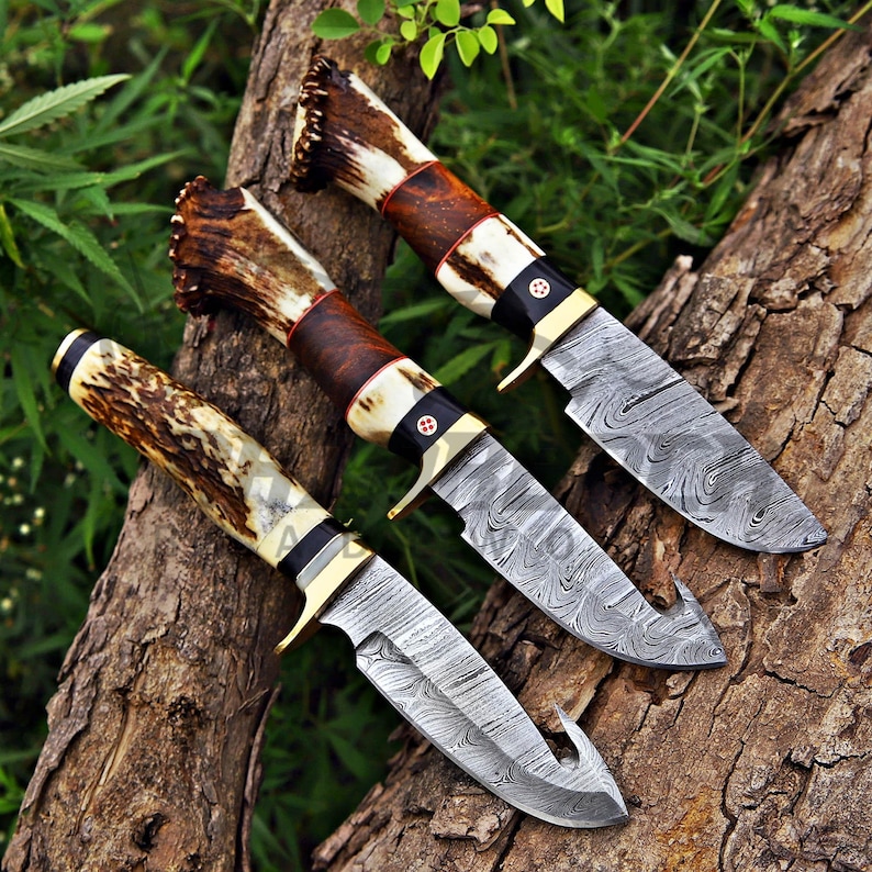 Custom Damascus hunting knife set with real stag antler handles, brass guards, and handcrafted steel blades displayed on rustic wood outdoors.