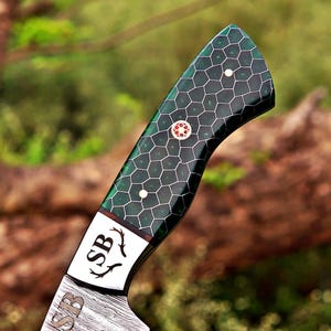 May include: A knife handle featuring a dark green honeycomb pattern with white hexagonal outlines. The handle includes a central circular mosaic pin and two smaller white pins. A metal bolster displays the letters SB with two antler icons.